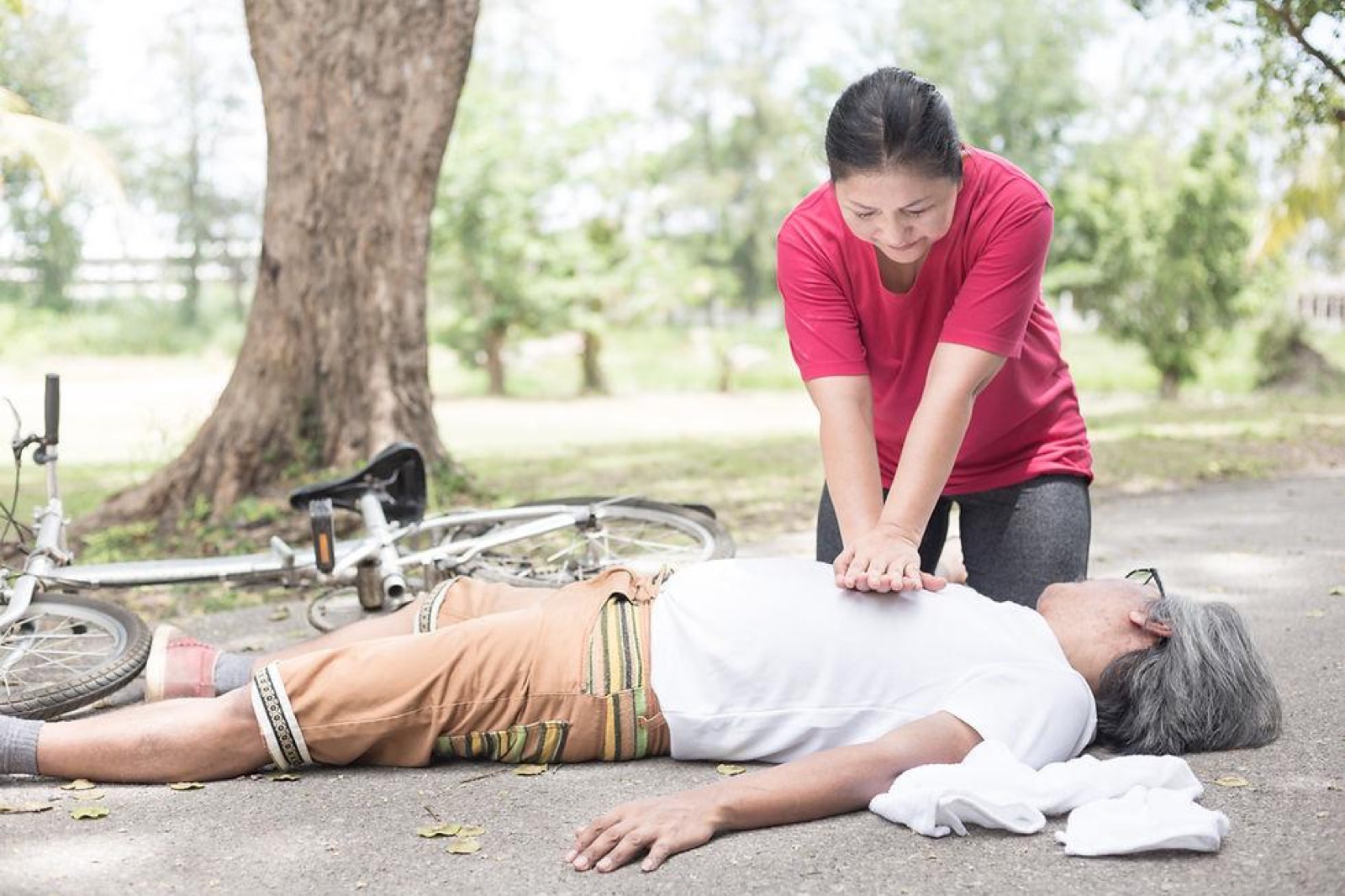 Une passante pratique les compressions thoraciques sur un cycliste
                effondré au bord d'un sentier.